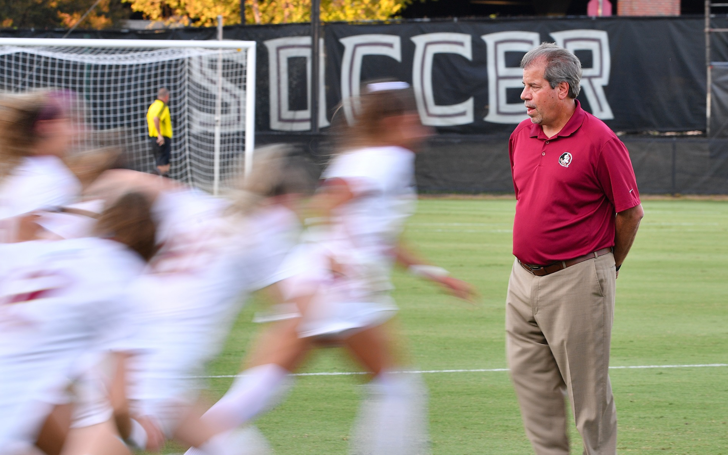 A photo of Coach Krikorian in a garnet polo, observing soccer players running on a field
