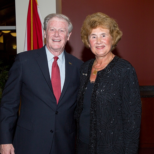 A photo of Joyce Miles wearing a black jacket, smiling, next to a man wearing a black suit