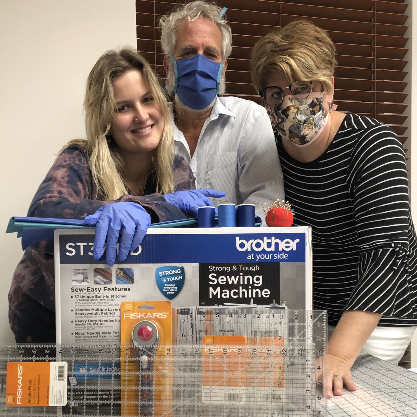 A photo of Annika Heetderks wearing blue surgical gloves, a man in a blue mask, and a woman in a patterned mask, standing in front of a boxed sewing machine
