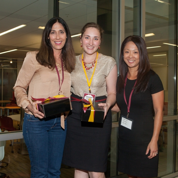 Three women wearing lanyards, smiling at the camera, two of whom are holding gift boxes