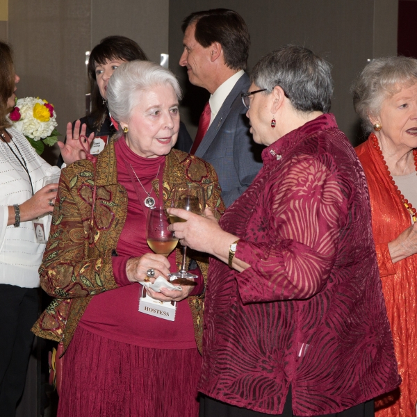 Two older women dressed in garnet and gold, holding glasses of white wine will speaking to each other