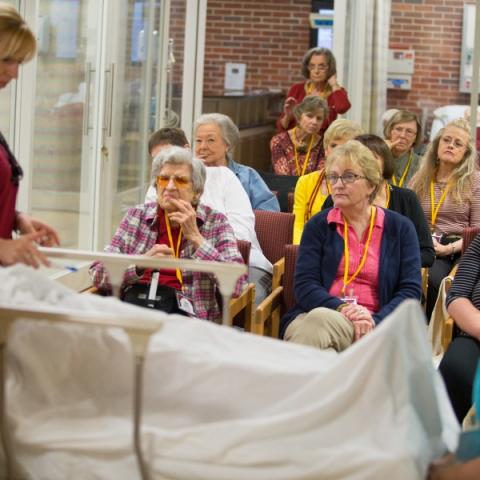 A group of older women watching someone give a presentation over a sheet-covered gurney