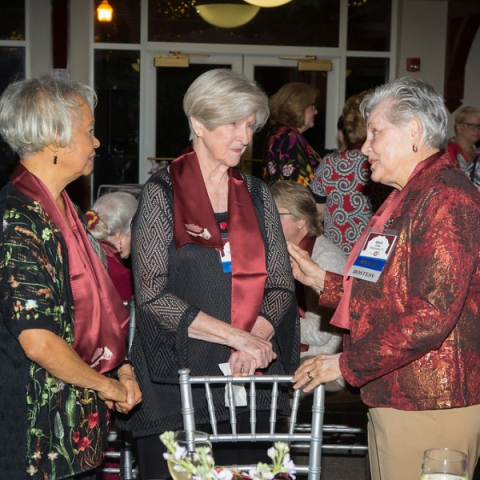 Three older women dressed in garnet having a conversation over a silver chair